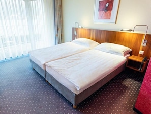 Una cama doble con cabecera de madera, una mesita de noche con lámpara y una ventana con cortinas traslúcidas.