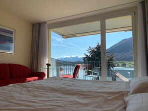 A hotel room with a bed, a red sofa, a chair, and a balcony with a mountain view.