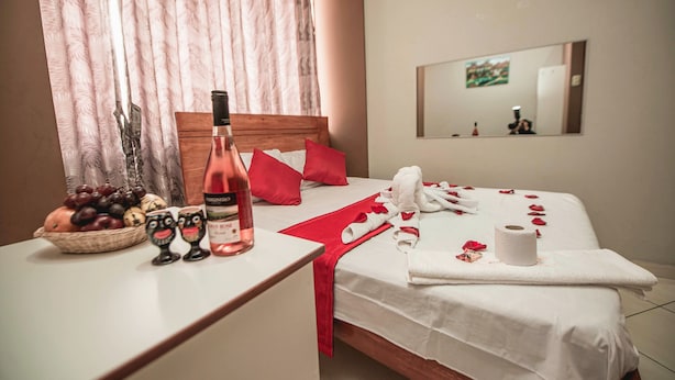 A neatly made bed with red pillows, a bottle of wine, and a bowl of fruit on the bedside table. - Hostal Spring II (Huánuco)