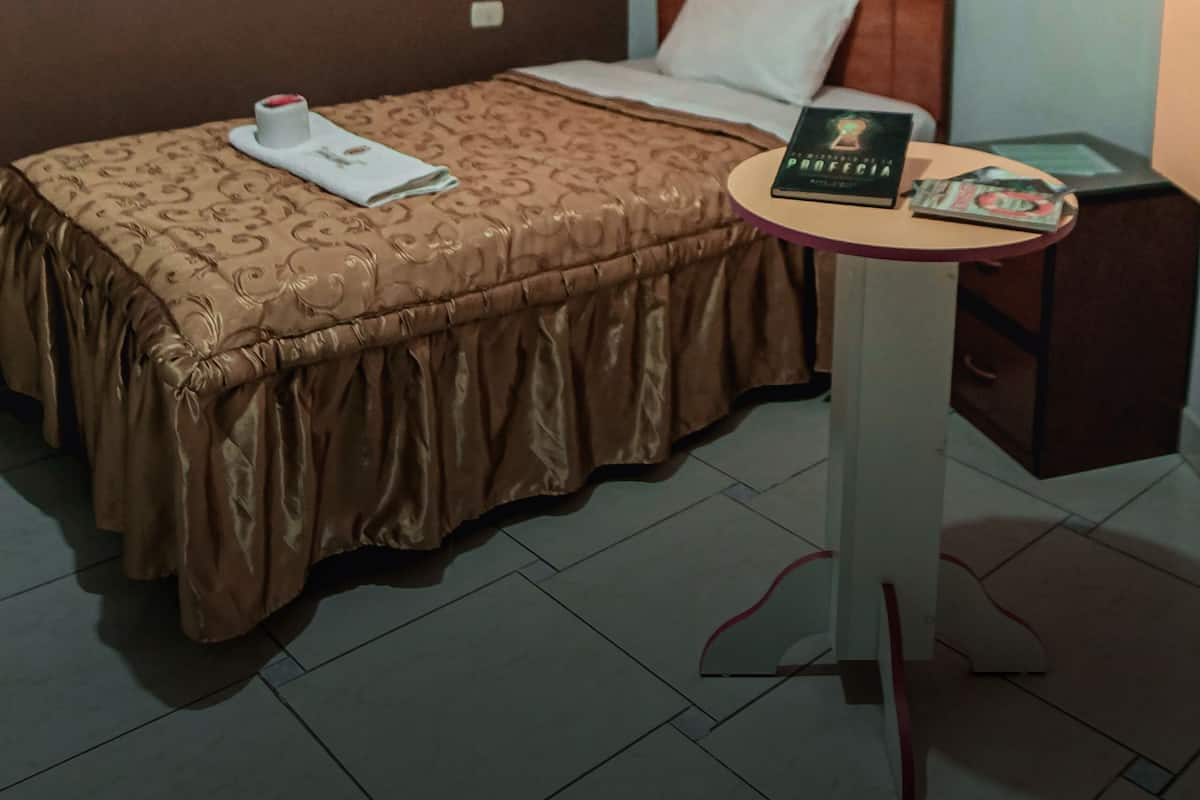 A neatly made bed with a brown bedspread and a white pillow, a small round table with a book and a magazine, and a wardrobe in the background.
