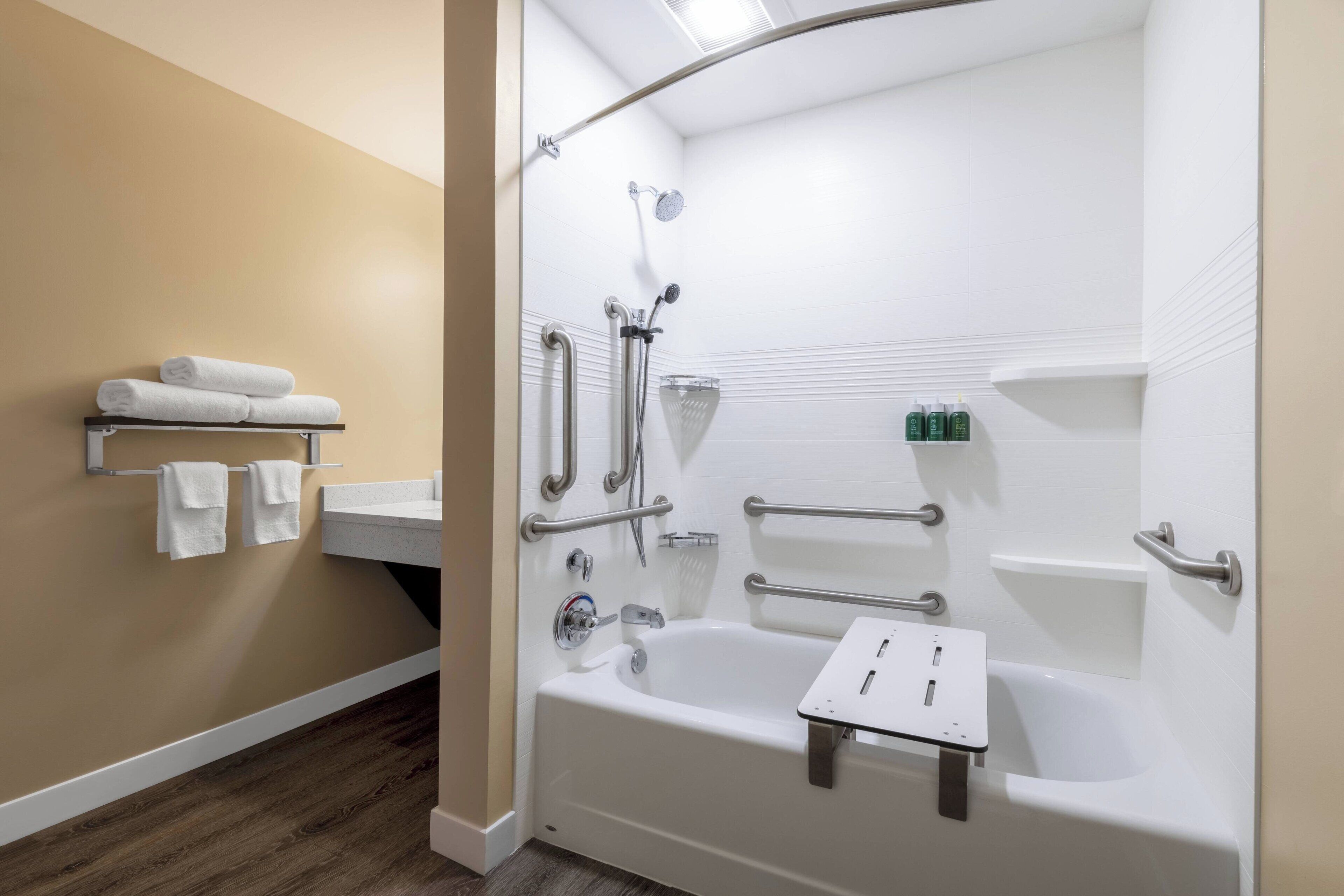 A modern bathroom with a bathtub, shower, and handrails. There are towels on a shelf and toiletries on a counter.