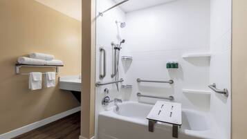 A modern bathroom with a bathtub, shower, and handrails. There are towels on a shelf and toiletries on a counter.