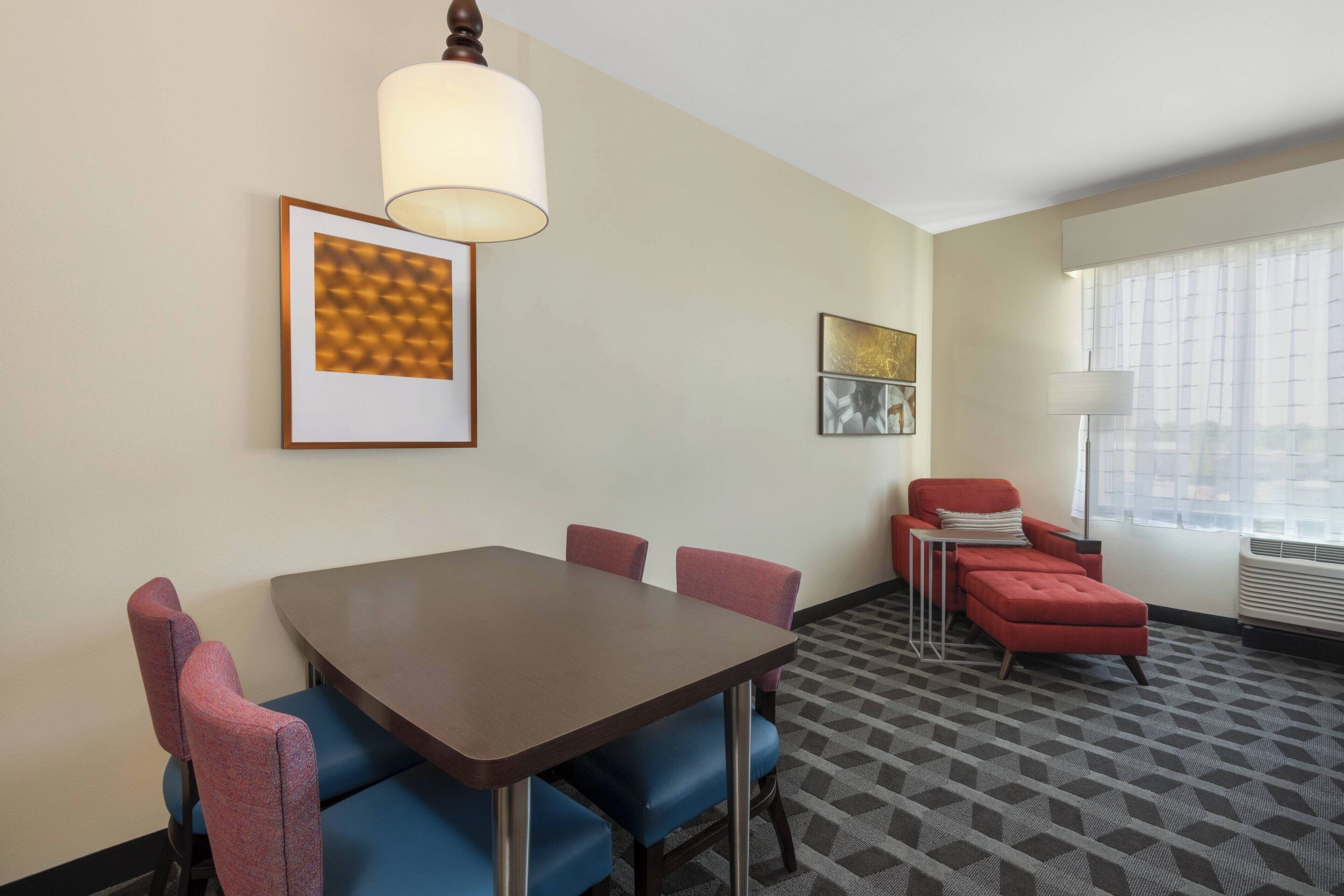 A hotel room with a dining table, chairs, a red sofa, and two framed artworks on the wall.