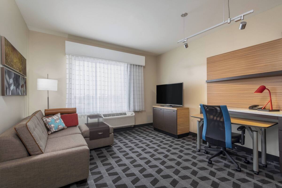 Una habitación de hotel con sofá, escritorio, televisión y ventana con cortinas.