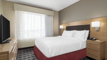 Habitación de hotel con una cama grande, un televisor en un aparador de madera, una mesita de noche con lámpara y una ventana con cortinas traslúcidas.