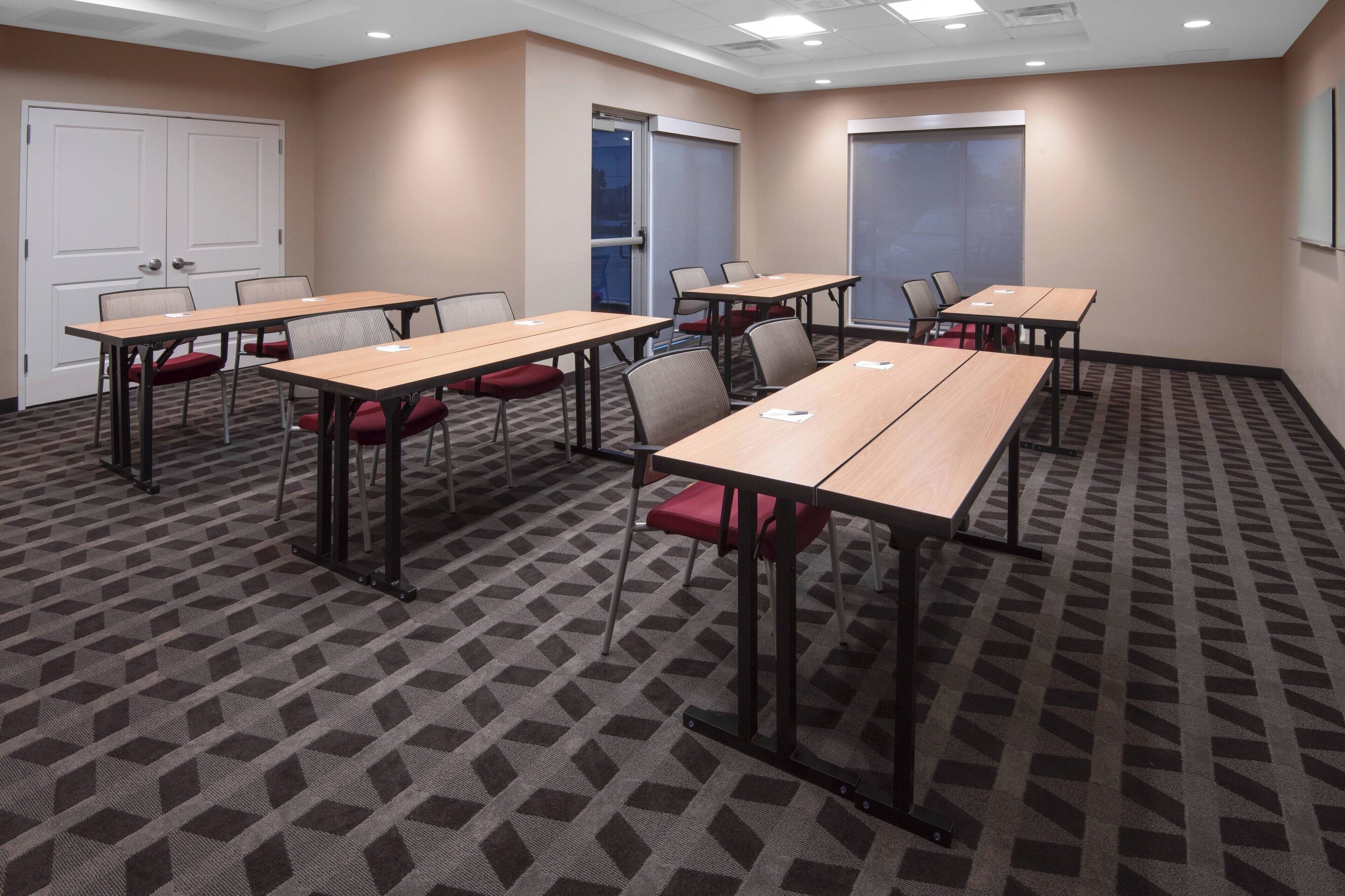 A conference room with six tables, each with four chairs. The room has a patterned carpet, beige walls, and a white door.