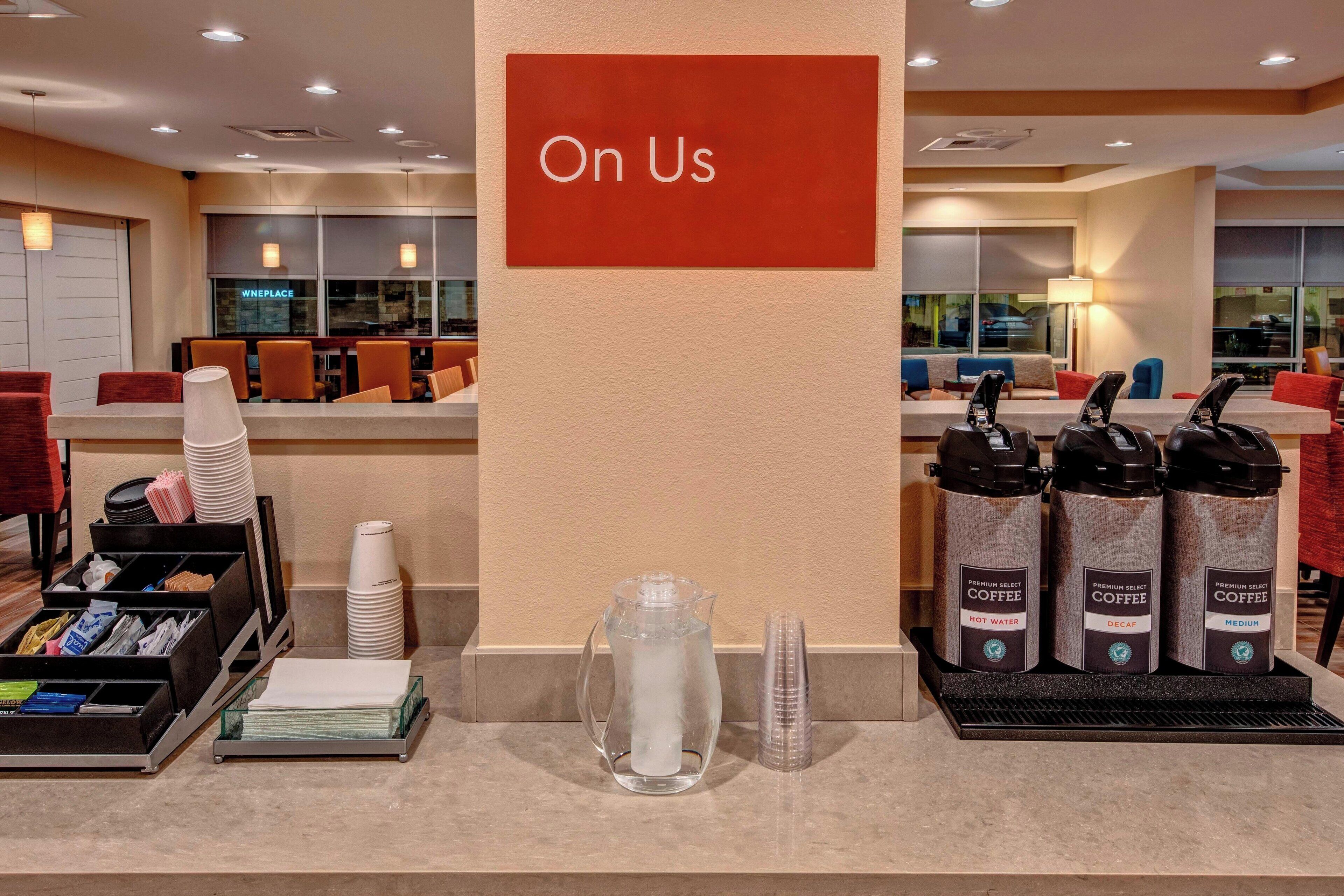 A hotel reception area with a 'On Us' sign, a coffee station with three coffee dispensers, and a water pitcher.