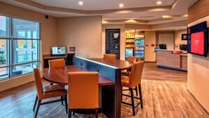 A hotel lobby with a wooden reception desk, a large dining table with chairs, a refrigerator stocked with beverages, and a digital screen displaying the hotel's name.