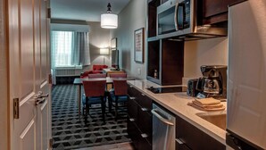 A compact hotel room with a kitchenette, a dining area with red chairs, a patterned floor, and a large window with curtains.