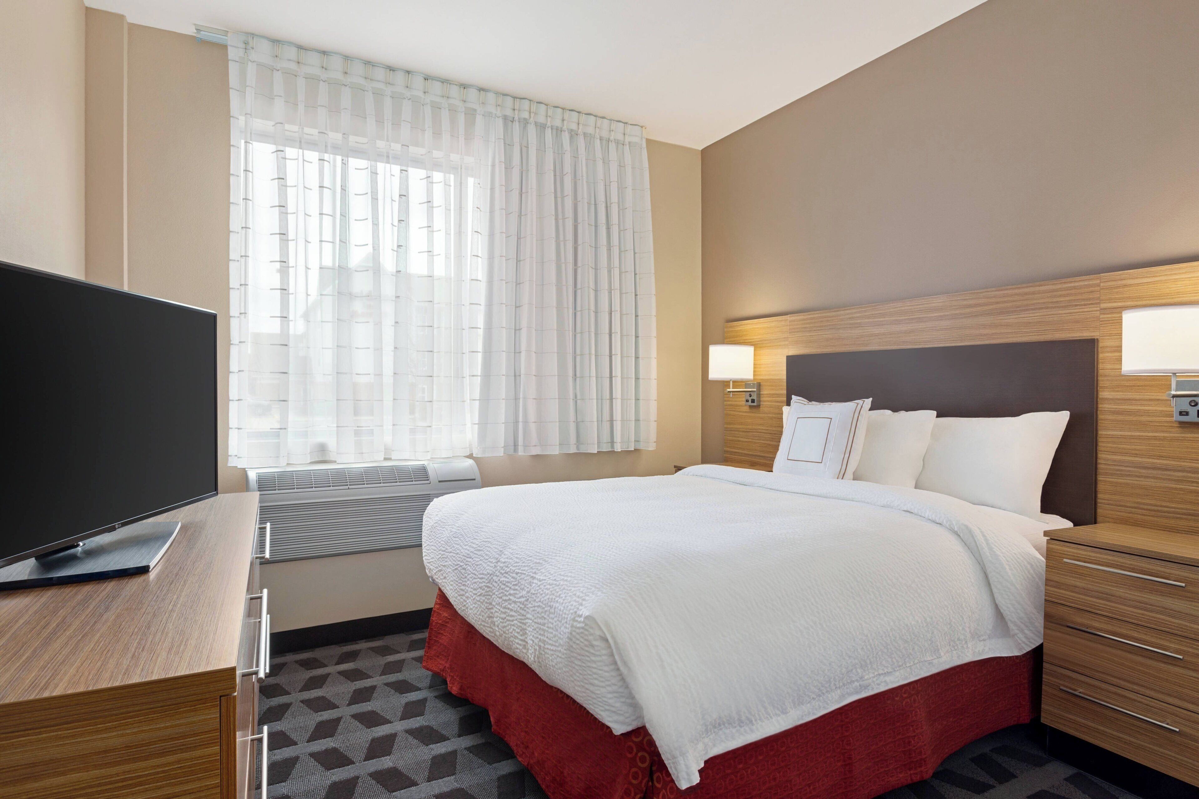 A hotel room with a large bed, a flat-screen TV, a bedside table, a window with sheer curtains, and a wall-mounted lamp.