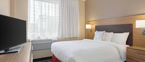 A hotel room with a large bed, a flat-screen TV, a bedside table, a window with sheer curtains, and a wall-mounted lamp.