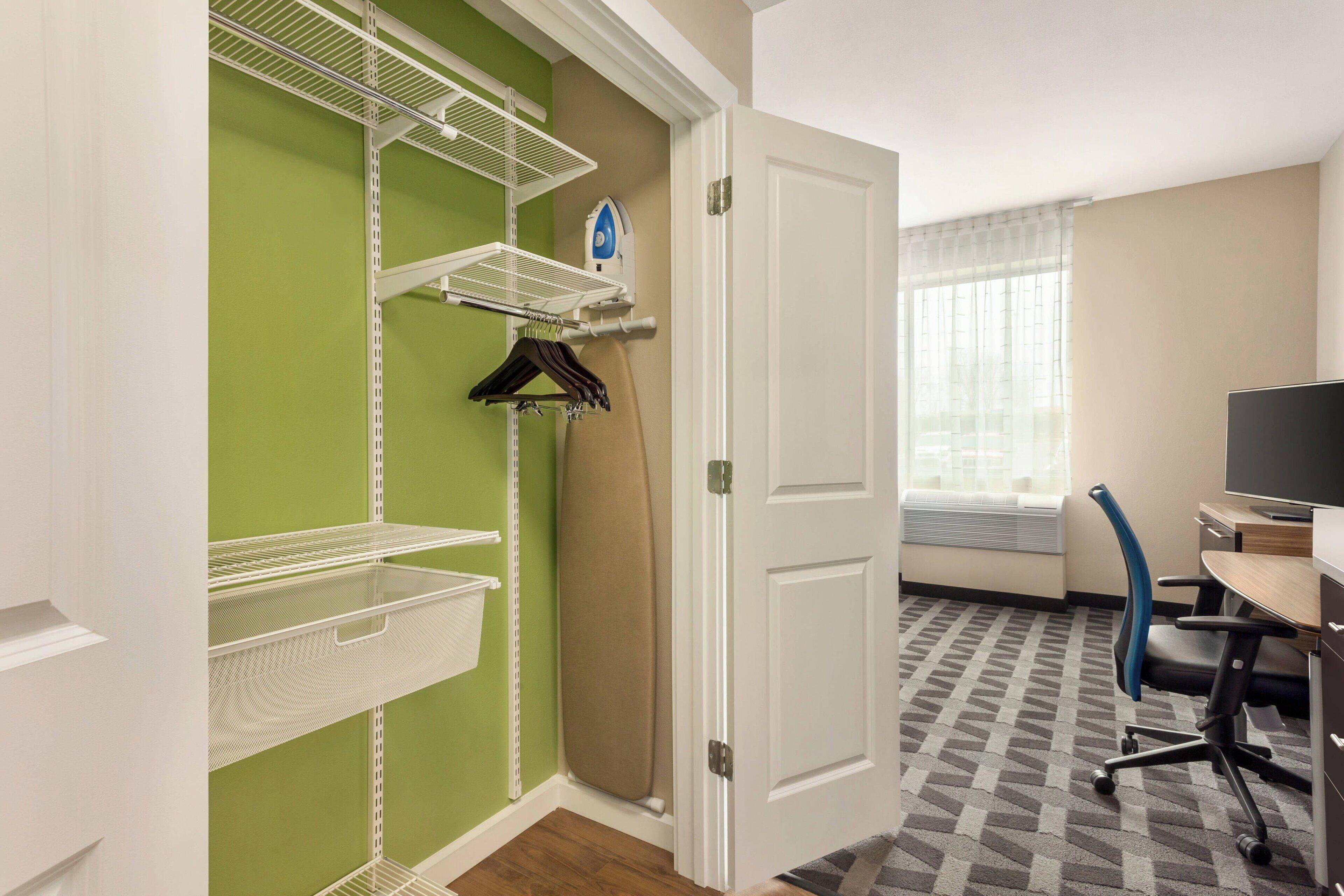 A hotel room with a green wall, white shelves, a desk with a computer, and a chair.