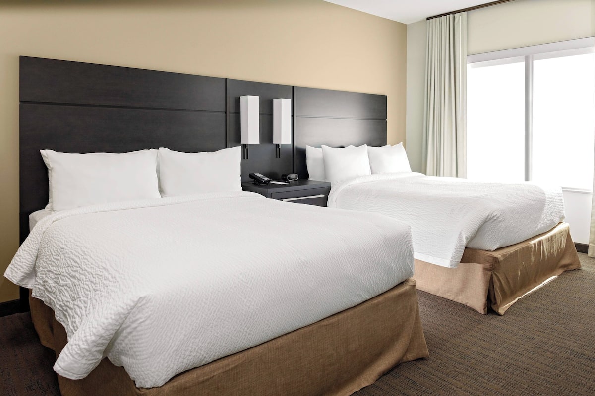 Ein Hotelzimmer mit zwei Betten, einem dunklen Kopfteil, Nachttischlampen, einem Nachttisch und einem großen Fenster mit Vorhängen.