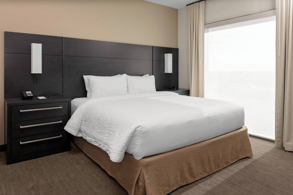 Ein Hotelzimmer mit einem großen Bett, einem Nachttisch mit Telefon und Blick ins Freie.