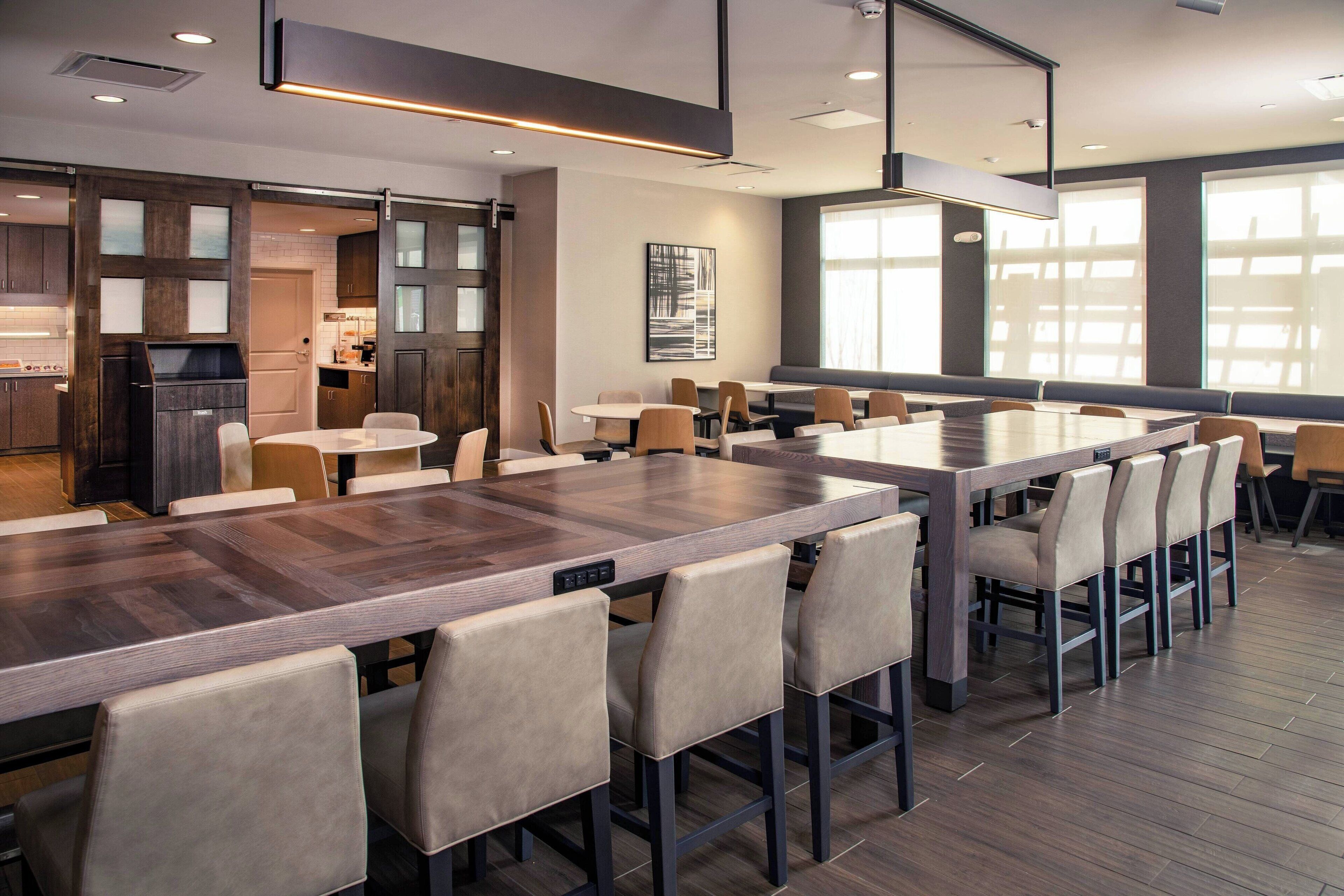 Uma área de jantar moderna com uma mesa de madeira comprida e várias cadeiras, uma cozinha com armários escuros e janelas amplas que proporcionam luz natural.