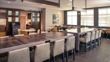 Uma área de jantar moderna com uma mesa de madeira comprida e várias cadeiras, uma cozinha com armários escuros e janelas amplas que proporcionam luz natural.
