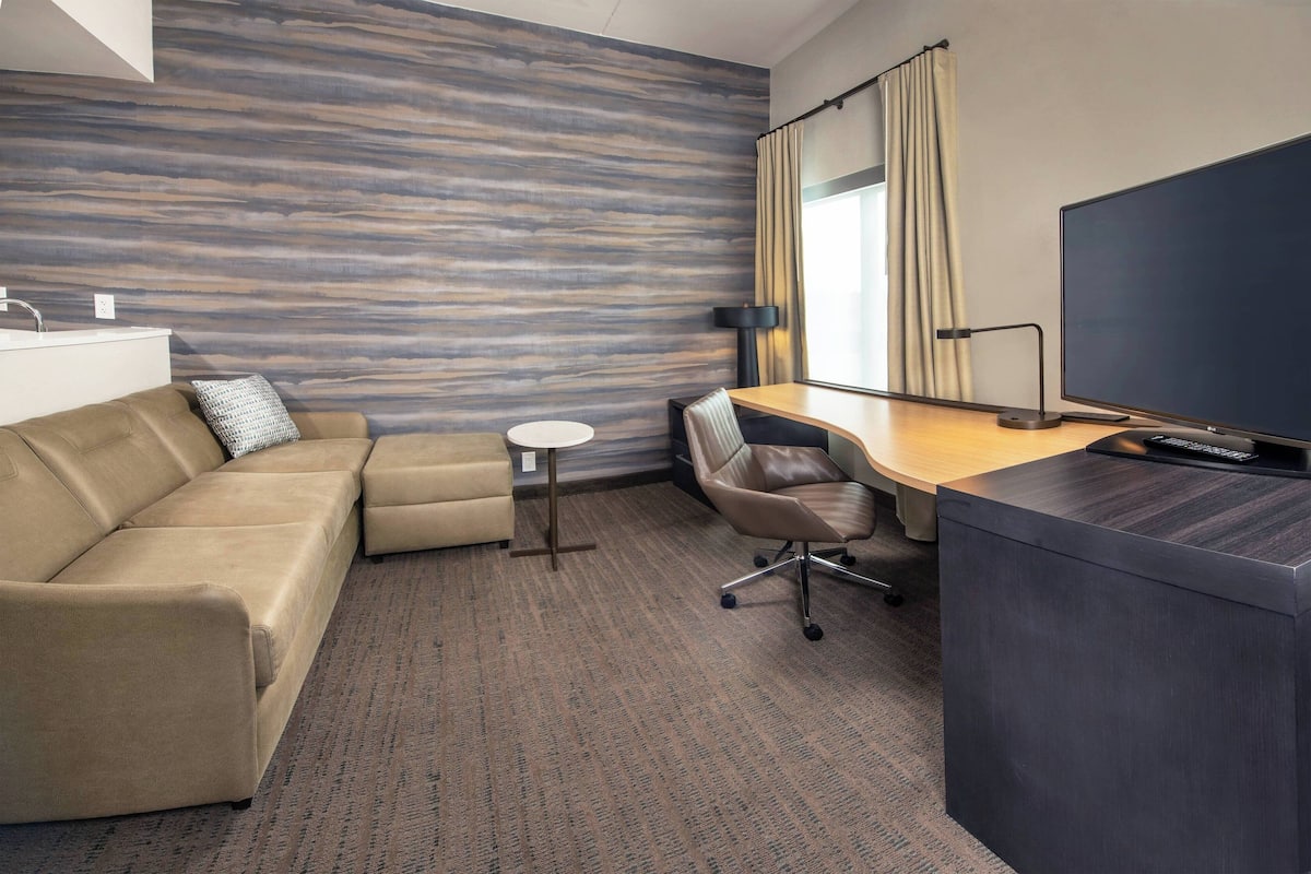 A hotel room with a sofa, a desk, a television, and a window with curtains.