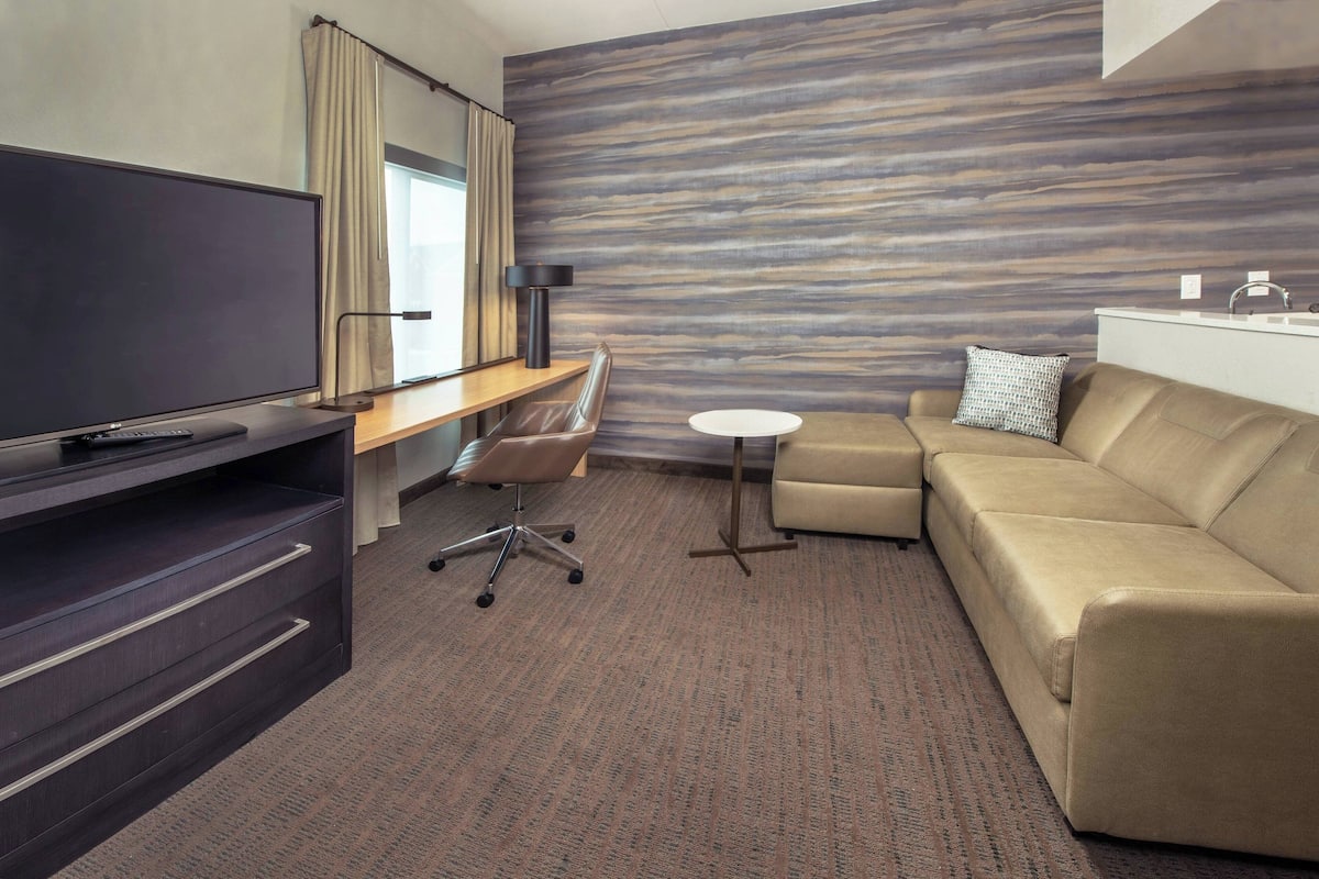 A hotel room with a flat-screen TV, a desk with a chair, a sofa, a small round table, and a wall with a wood-like pattern.