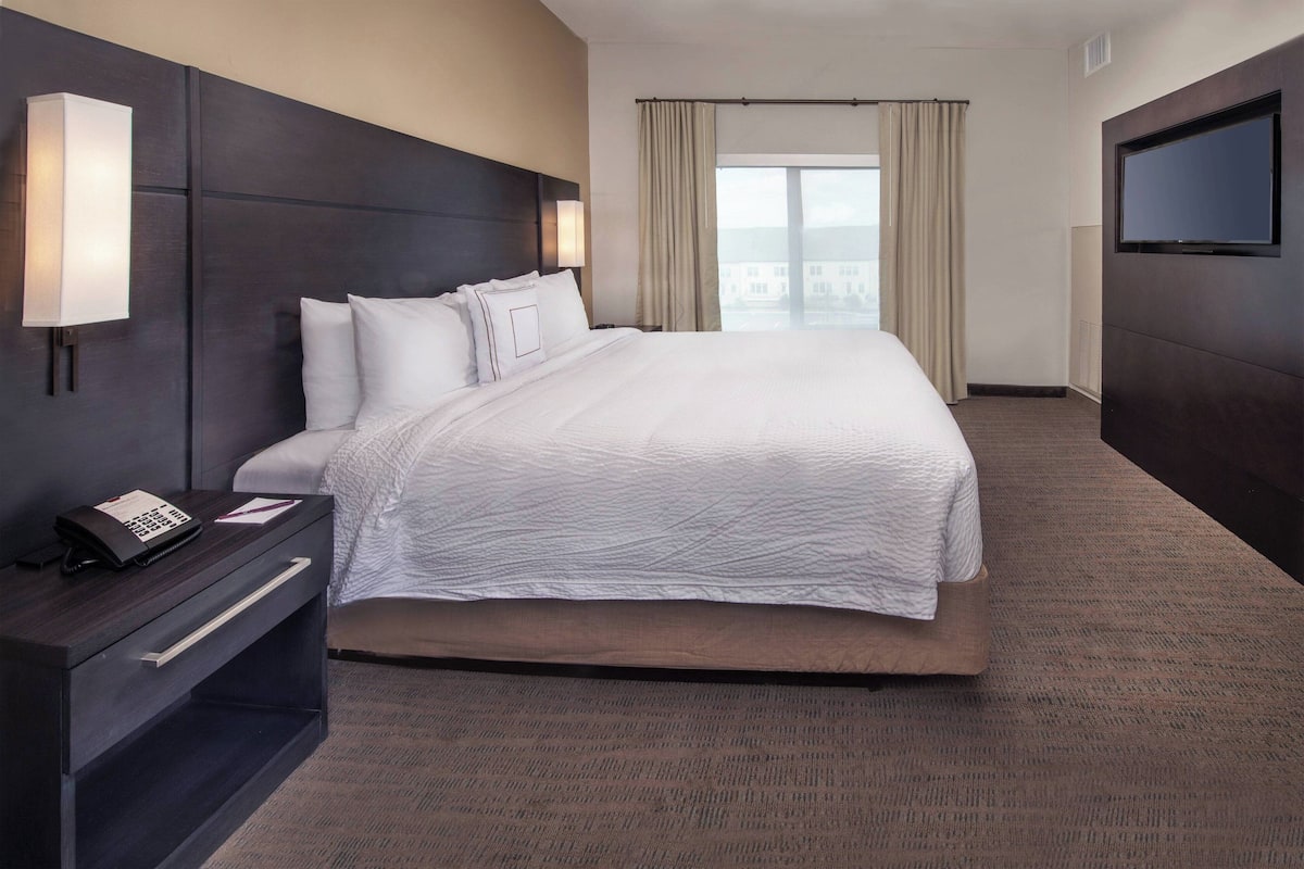 A hotel room with a large bed, a bedside table with a phone, a flat-screen TV mounted on the wall, and a view of the city through the window.