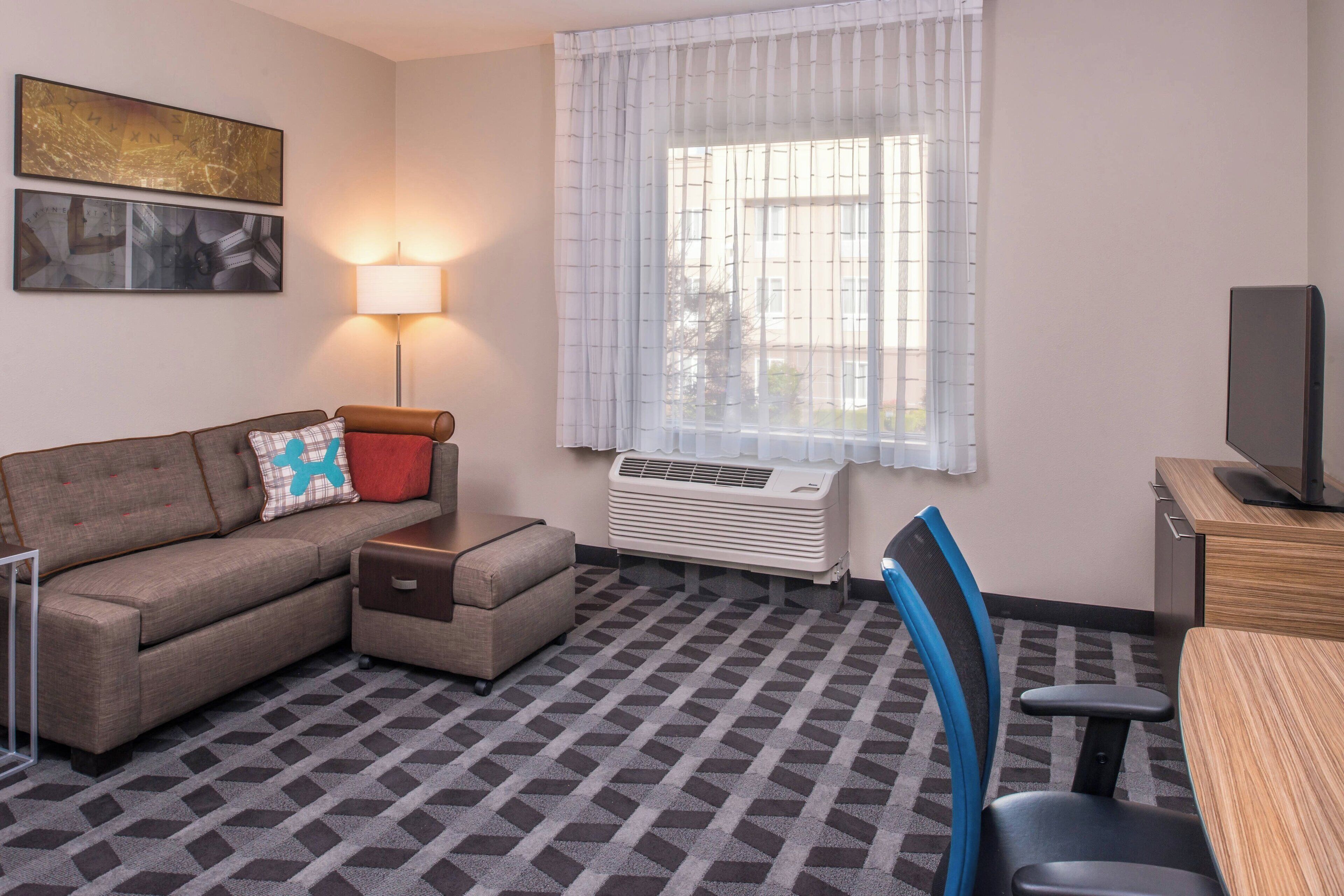 A hotel room with a sofa, a small table, a chair, a TV, a lamp, and a window with curtains.