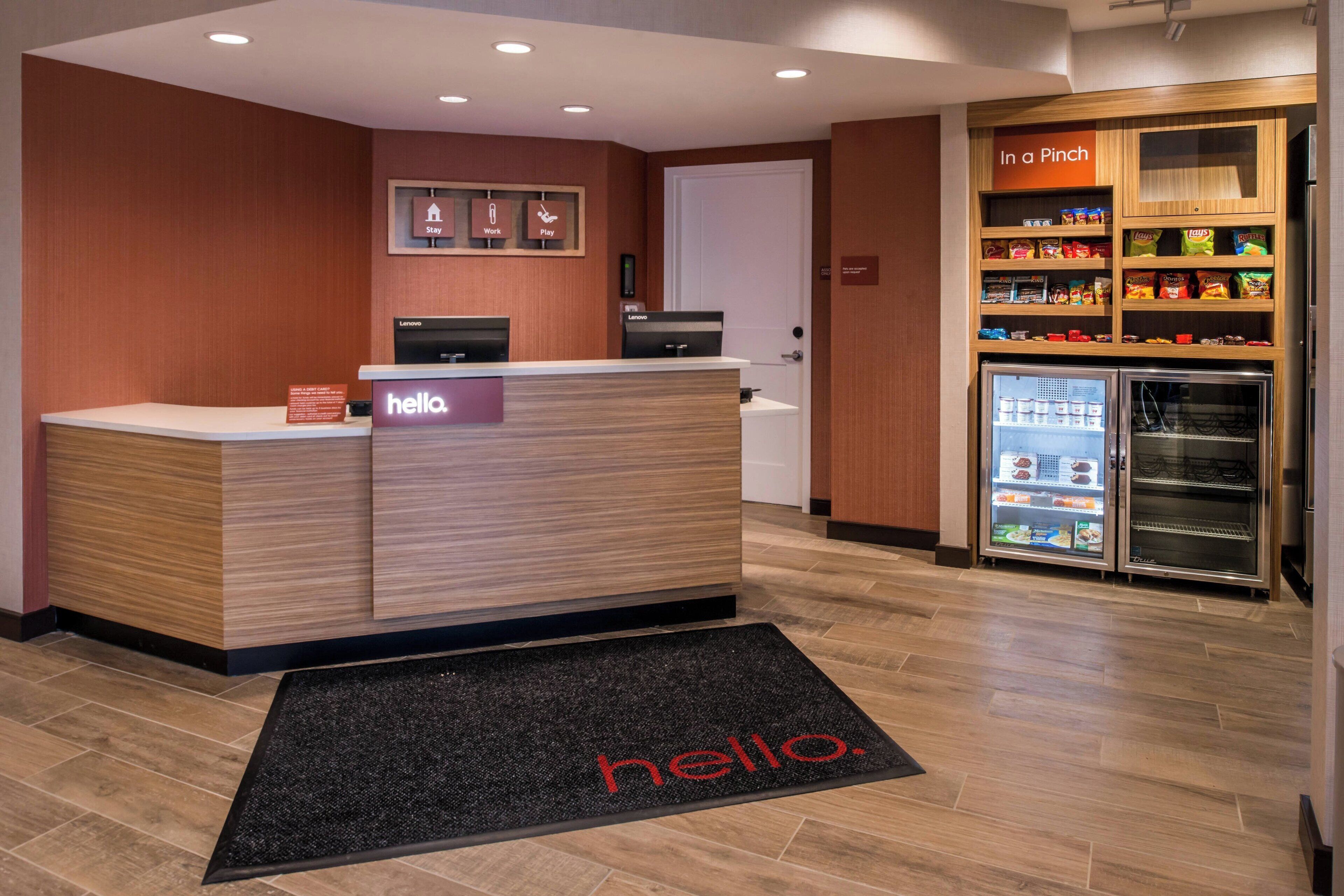 Ein Hotel-Rezeptionsbereich mit Empfangstresen, einer Fußmatte mit der Aufschrift 'hello', einem mit Snacks befüllten Kühlschrank und einem Regal mit der Bezeichnung 'In a Pinch', voll mit verschiedenen Artikeln.