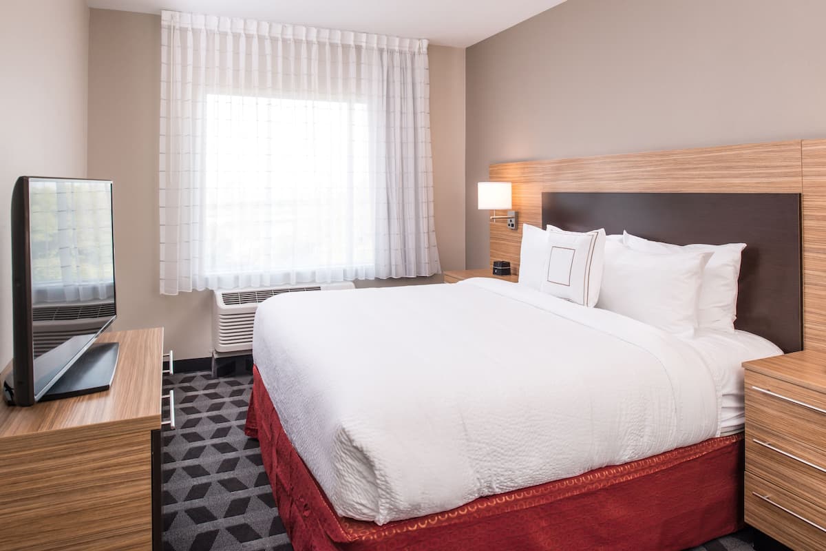 A hotel room with a large bed, a TV on a wooden desk, a window with sheer curtains, and a bedside lamp.