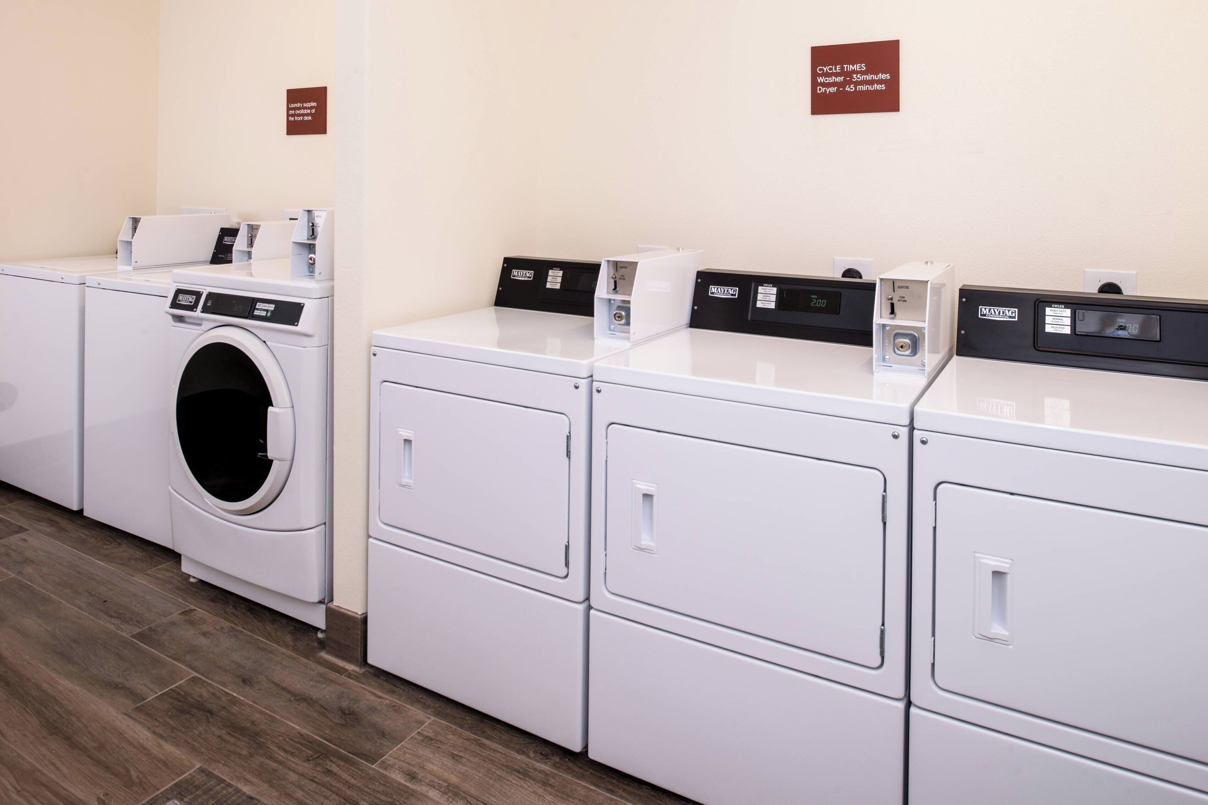 A laundry facility with a front-loading washer and dryers, a sign indicating cycle times, and a wooden floor.