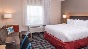 A hotel room with a large bed, a desk with a chair, a sofa with a pillow, a window with curtains, and an air conditioning unit.