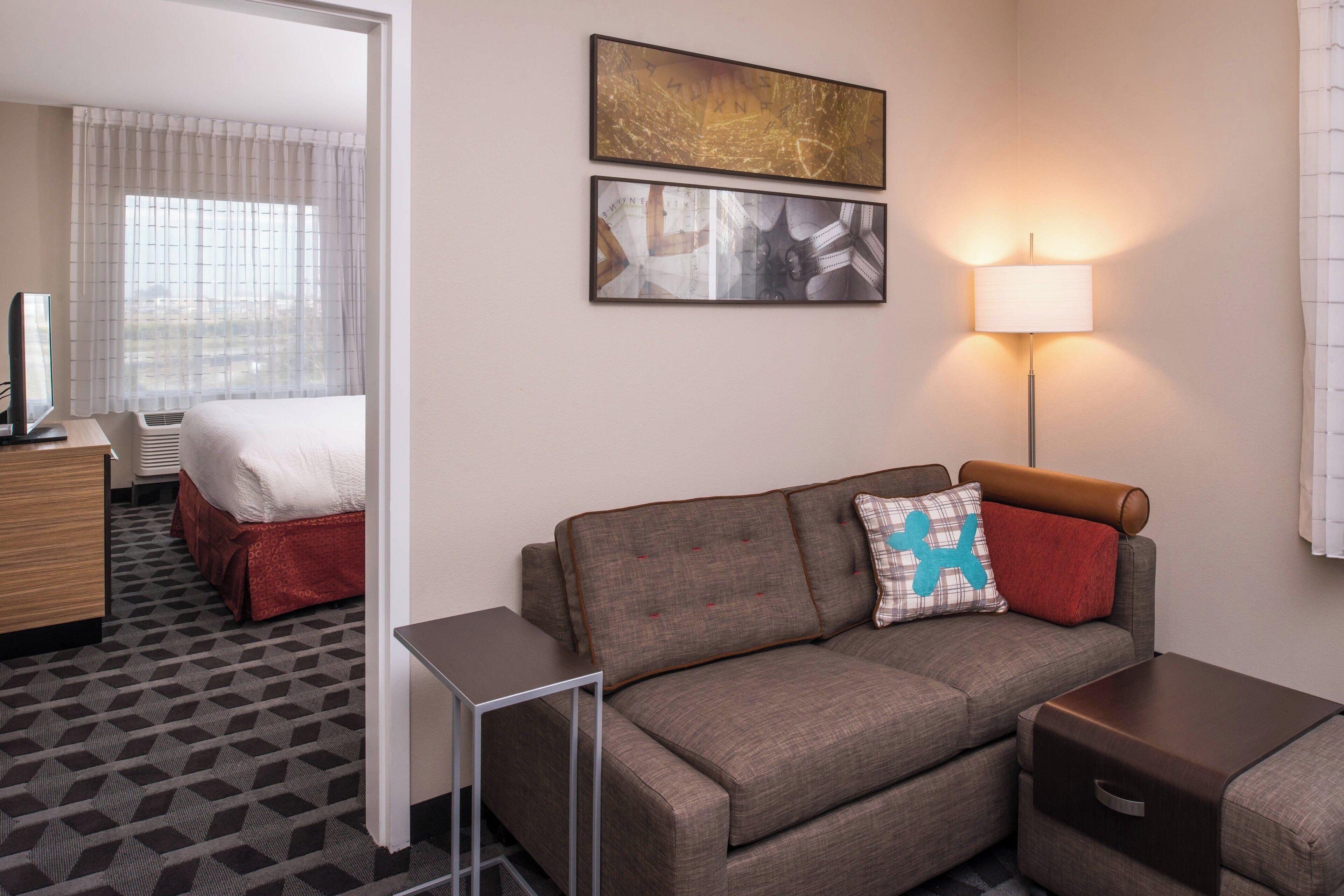 A hotel room with a sofa, a lamp, a small table, and a bed with a red bed skirt.