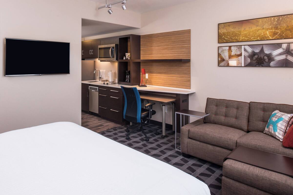 A modern hotel room with a bed, a sofa, a desk, and a kitchenette.
