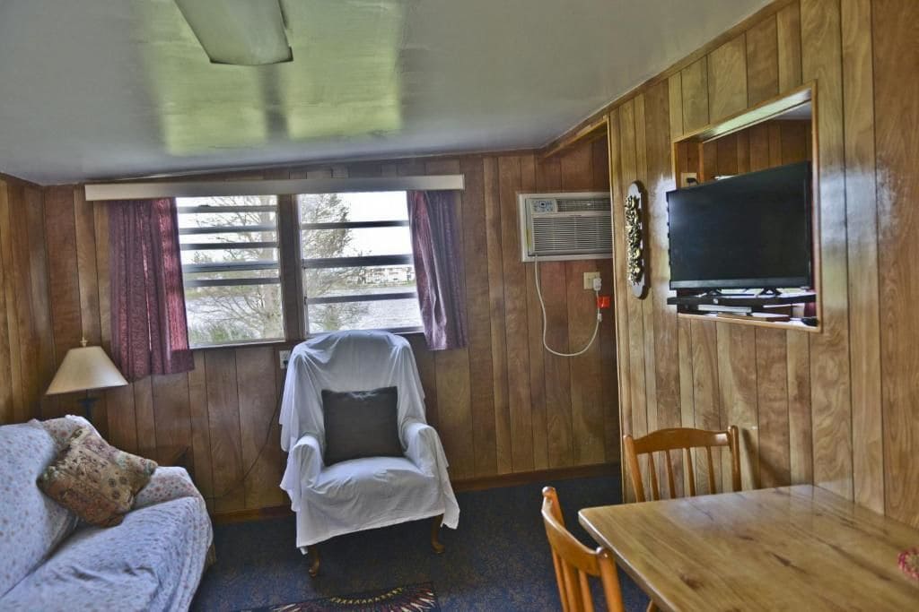 A room with wooden walls, a television mounted on a wooden panel, a covered chair, a wooden table, and a window with curtains.