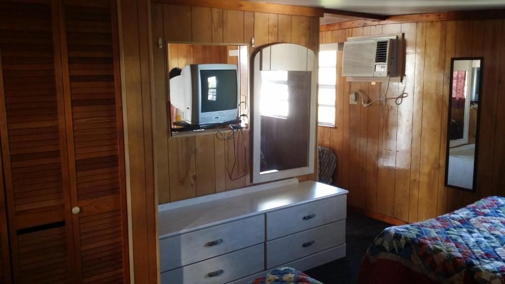 A bedroom with wooden paneling, a dresser with drawers, a bed with a quilt, a television on a stand, and an air conditioning unit.