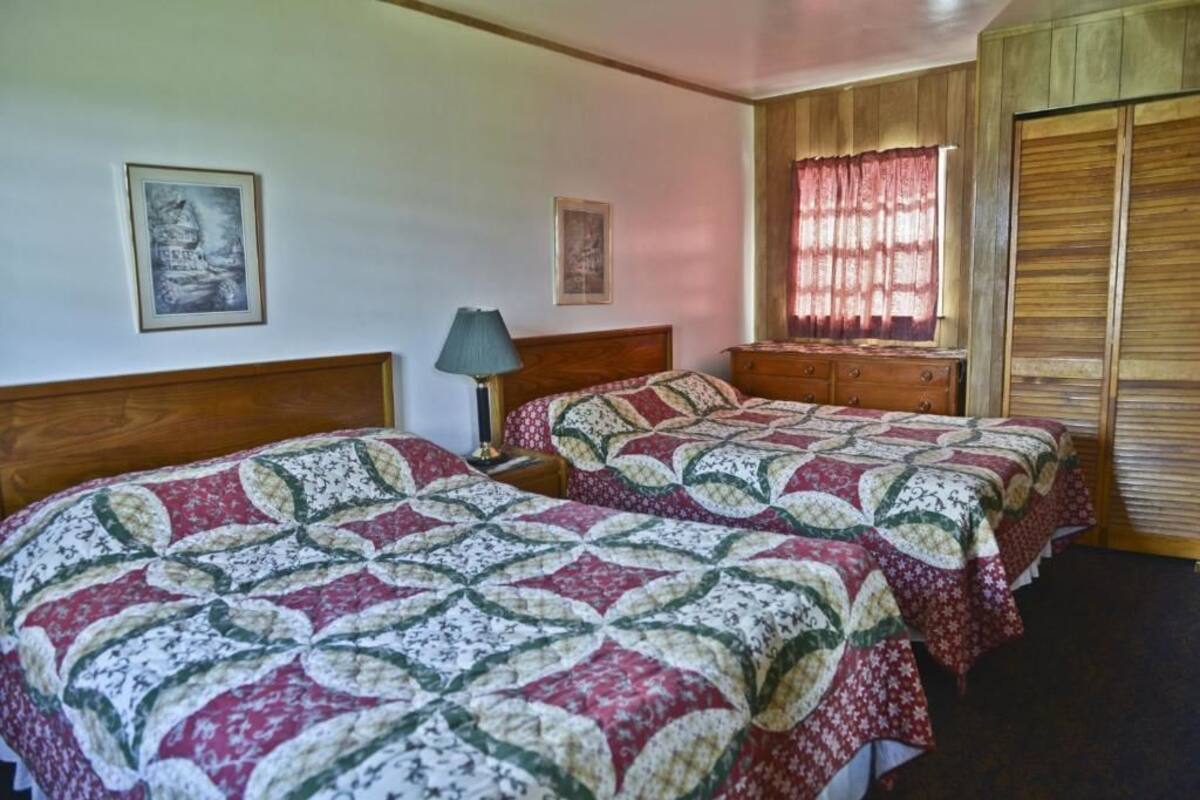 A bedroom with two beds, wooden headboards, a nightstand, a lamp, and a window with curtains.