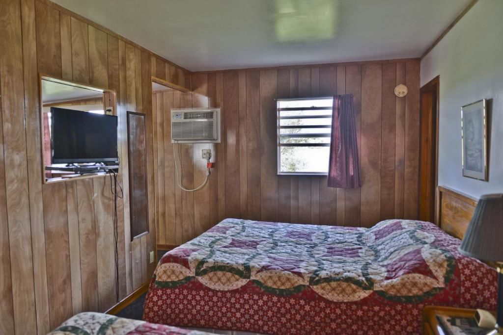 A room with wooden paneling, a bed with a quilt, a television, an air conditioning unit, and a window with blinds.