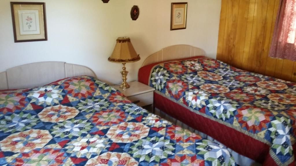A room with two beds, each with a quilted bedspread. There is a bedside table with a lamp between the beds.