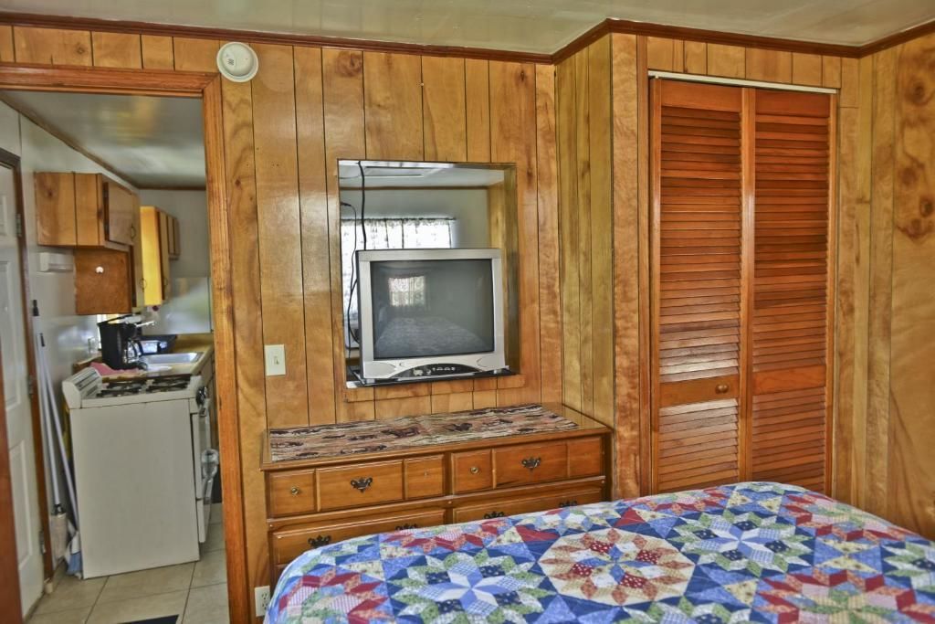 A room with wooden paneling, a bed with a quilt, a television on a dresser, and a kitchenette with a stove and cabinets.