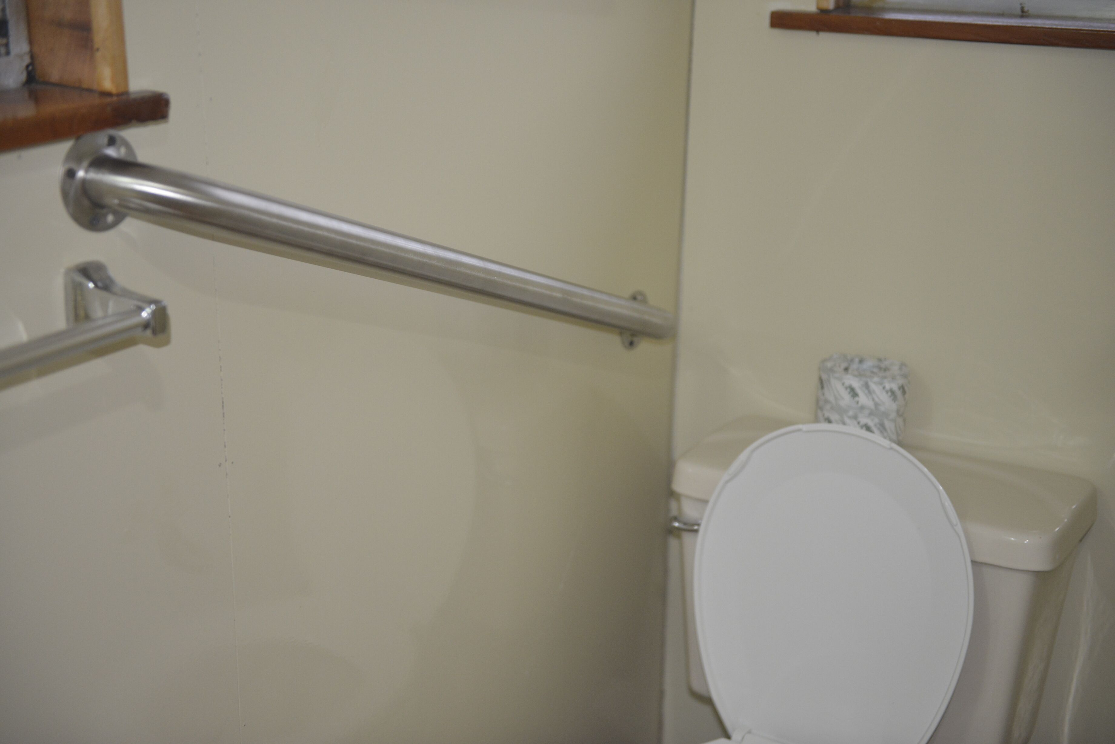 A bathroom with a white toilet, a metal grab bar, and a wooden shelf above the toilet.