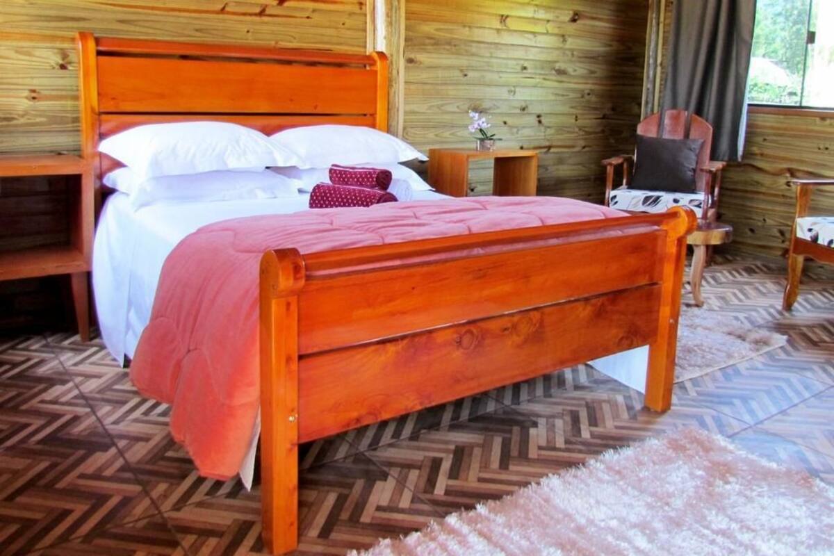 Una cama de madera con cabecera, una mesita de noche, una silla y una pequeña mesa con un florero.
