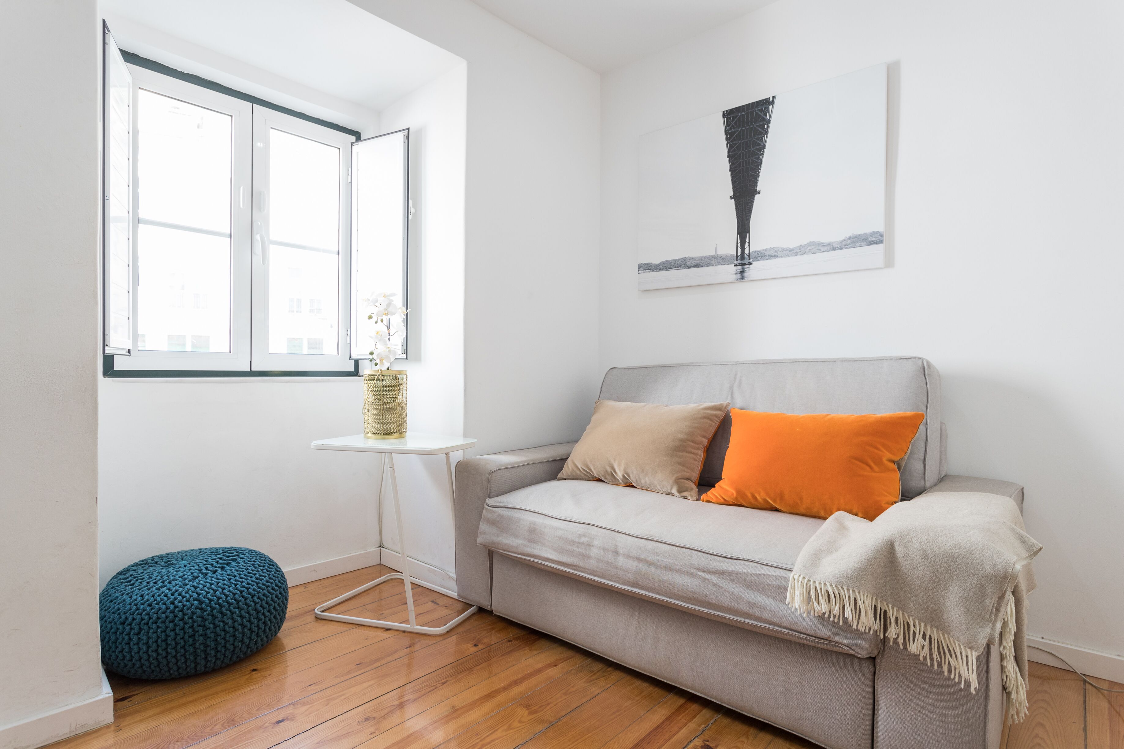 A modern living room with a grey sofa, orange cushions, a white throw blanket, a small white side table with a vase, and a blue ottoman.