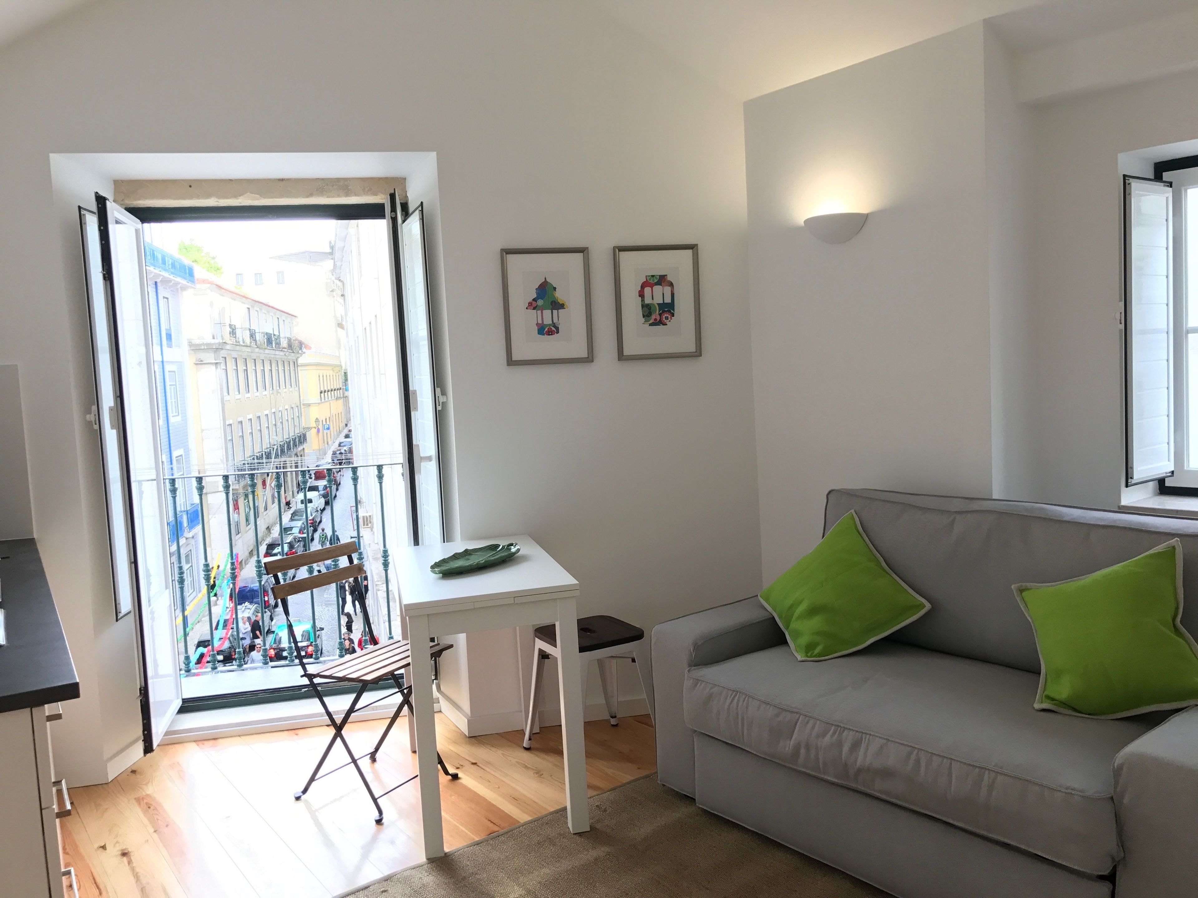 A modern living room with a grey sofa, green cushions, a small white table, and a balcony with a view of buildings and parked cars.