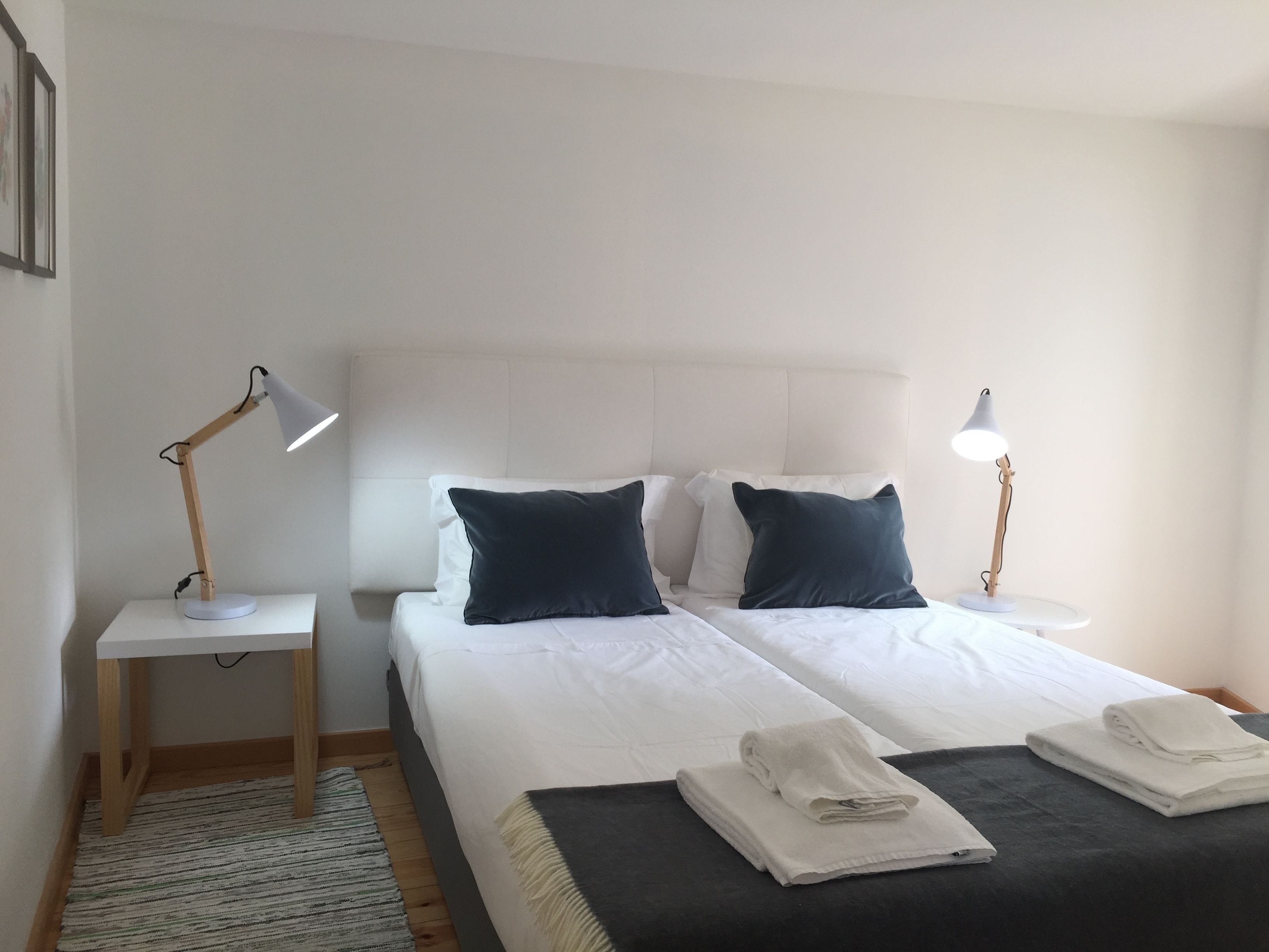 A hotel room with two beds, white bedding, dark pillows, a white headboard, two bedside tables with lamps, and a folded towel on each bed.