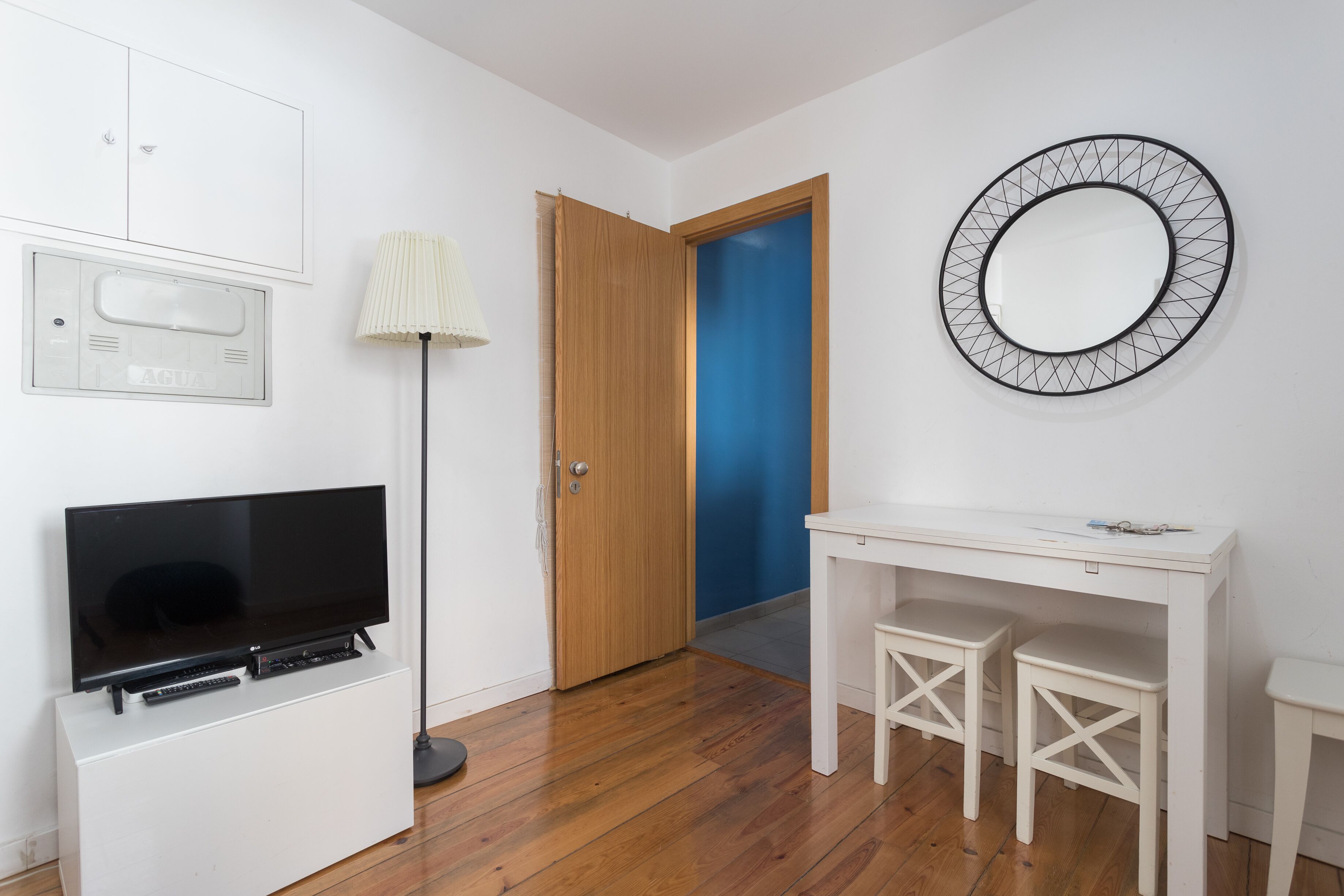 A room with a flat-screen TV on a white cabinet, a wooden door, a blue wall, a white desk with two stools, and a wall-mounted mirror.