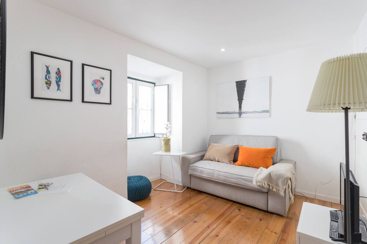 Ein modernes Wohnzimmer mit einem grauen Sofa, einem weißen Couchtisch und einer Lampe. An der Wand befinden sich gerahmte Kunstwerke und ein Fenster sorgt für natürliches Licht.