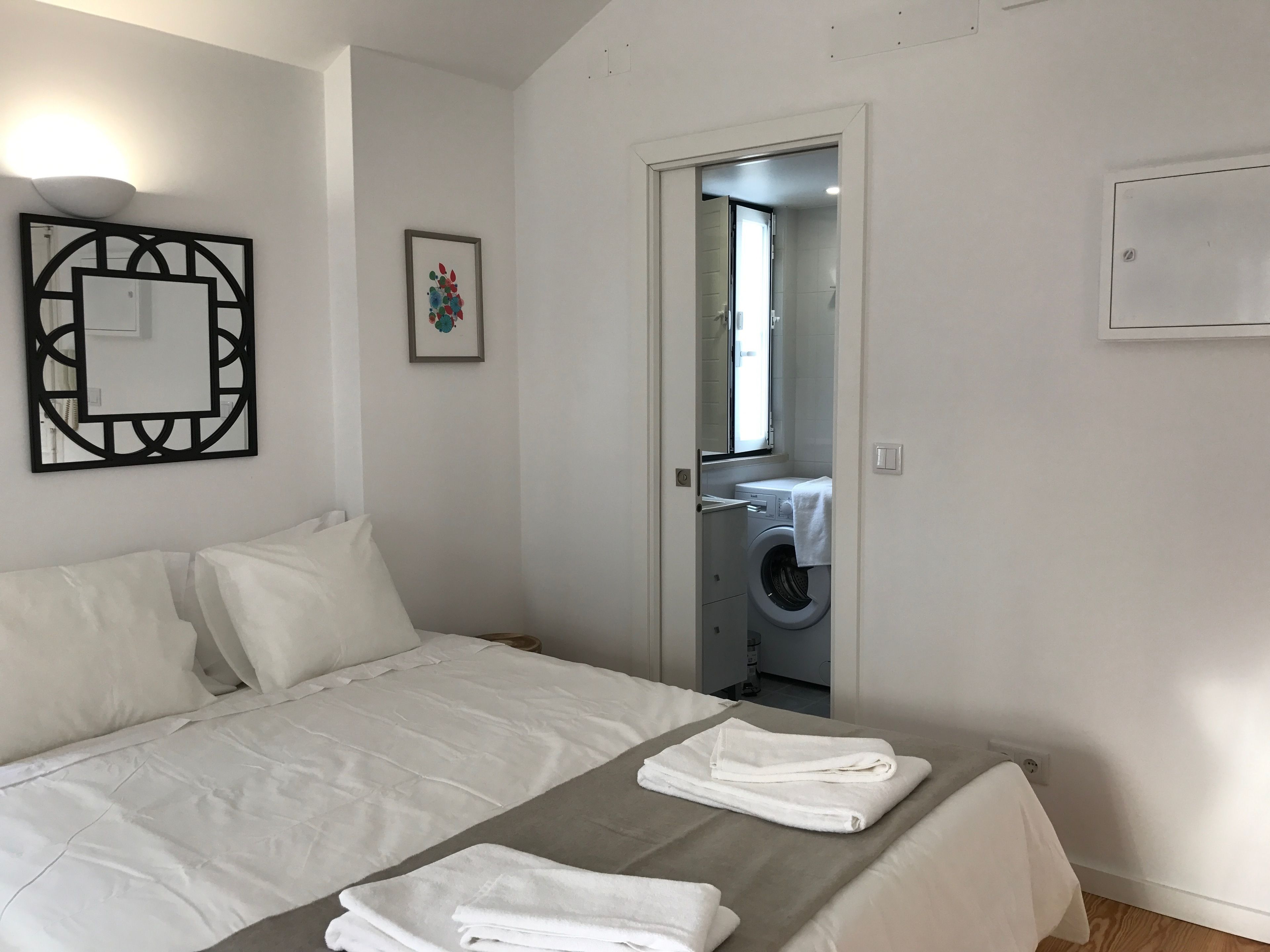 Un dormitorio con cama, una mesita de noche con toallas, un espejo montado en la pared y una puerta que da a un baño con lavadora.