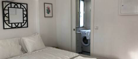 Un dormitorio con cama, una mesita de noche con toallas, un espejo montado en la pared y una puerta que da a un baño con lavadora.