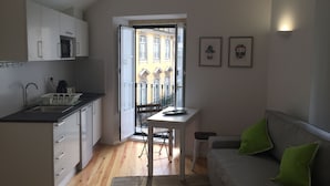 Une cuisine moderne avec des meubles blancs, un four à micro-ondes et un évier. On y trouve une table à manger avec des chaises, un canapé avec des coussins verts et un balcon avec vue sur les immeubles.