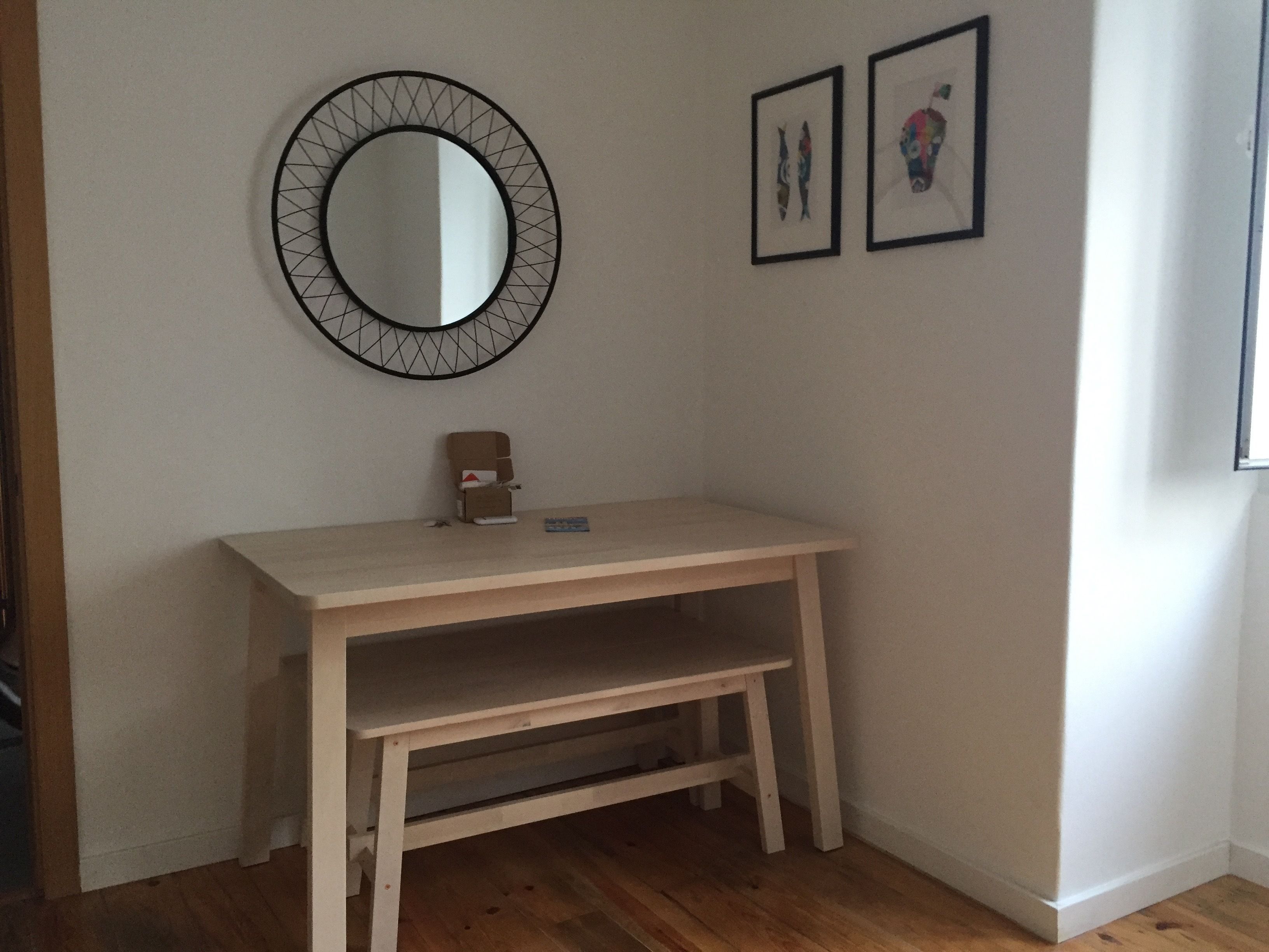 Quarto com escrivaninha de madeira, espelho redondo com moldura geométrica e duas obras de arte emolduradas na parede.