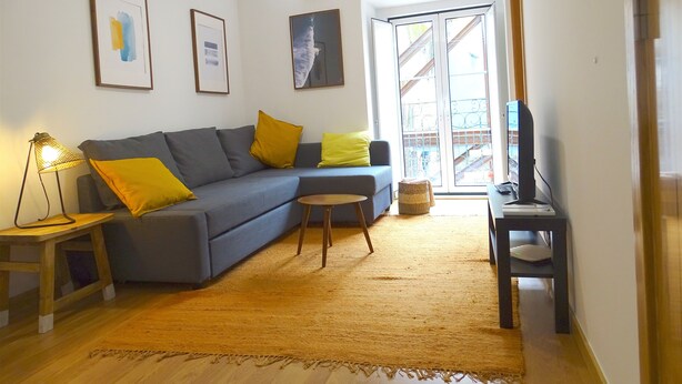 Ein Wohnzimmer mit grauem Sofa, gelben Kissen, einem kleinen Holztisch, einer Stehlampe und einem Fernseher auf einem Schrank.