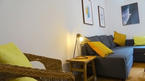 Ein Wohnzimmer mit einem Korbstuhl, einem kleinen Holzschemel, einem blauen Sofa mit gelben Kissen und einer Stehlampe.