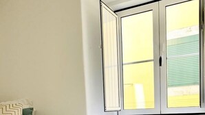 A bedroom with two beds, a window with blinds, a bedside table with a lamp, and a woven basket on the floor.
